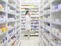 Farmaceutka w białym fartuchu idzie między regałami pełnymi leków w aptece.