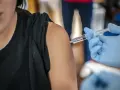 Osoba w czarnej koszulce otrzymuje zastrzyk. Ręka w niebieskiej rękawiczce trzyma strzykawkę, gotową do podania szczepionki, być może chroniącej przed varilrix.