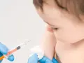 Dłoń w niebieskiej rękawiczce trzyma strzykawkę, przygotowując się do szczepienia niemowlęcia.