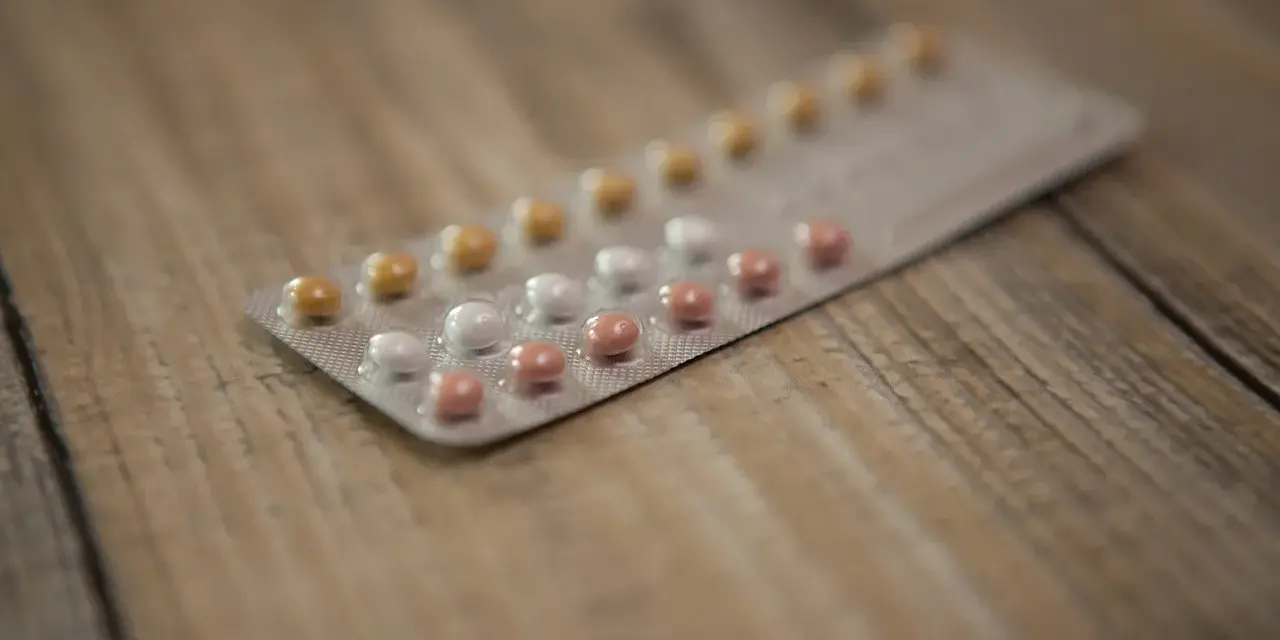 Opakowanie tabletek antykoncepcyjnych z białymi, różowymi i żółtymi pigułkami na drewnianym tle.
