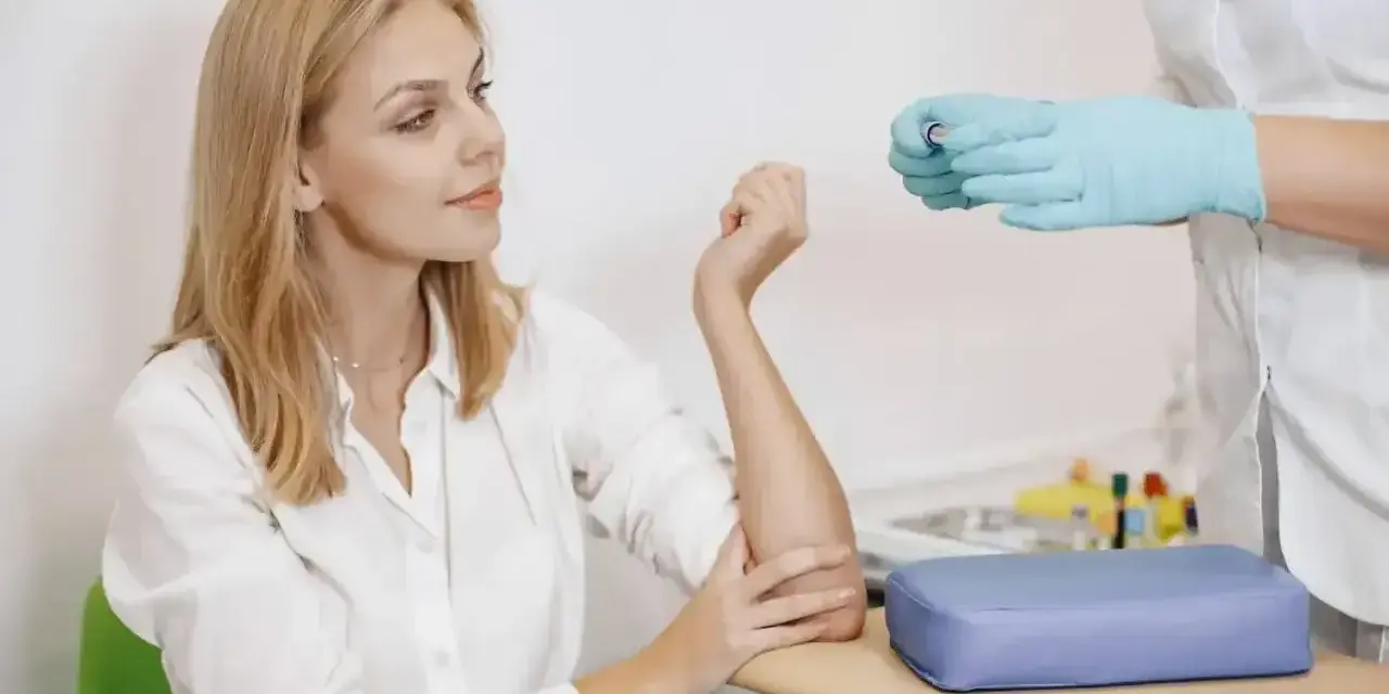 Kobieta w białej koszuli przygotowuje się do pobrania krwi. Pielęgniarka w rękawiczkach trzyma probówkę. To standardowa procedura, gdy badanie wykonuje się na czczo.