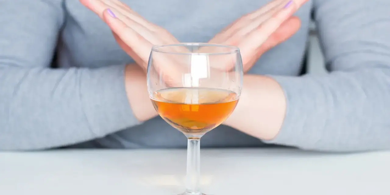 Ręce w geście "stop" nad kieliszkiem z bursztynowym płynem. Pytanie: czy po szczepieniu można pić alkohol? Odpowiedź brzmi: nie.