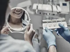 Dentysta pokazuje pacjentce wzornik kolorów zębów, pacjentka ogląda swój uśmiech w lusterku.