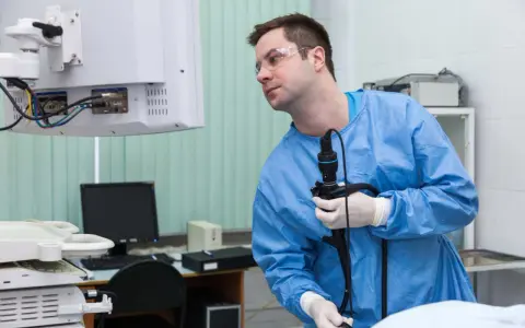 Lekarz w niebieskim fartuchu i okularach ochronnych trzyma endoskop, patrząc na monitor.