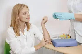 Kobieta w białej koszuli przygotowuje się do pobrania krwi. Pielęgniarka w rękawiczkach trzyma probówkę. To standardowa procedura, gdy badanie wykonuje się na czczo.